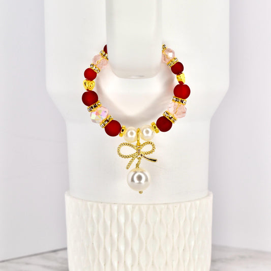 A beaded tumbler handle bracelet with red, pink, and gold accents. The bracelet features a gold bow charm with a dangling pearl. The bracelet is displayed on a white Stanley.