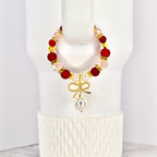 A beaded tumbler handle bracelet with red, pink, and gold accents. The bracelet features a gold bow charm with a dangling pearl. The bracelet is displayed on a white Stanley.