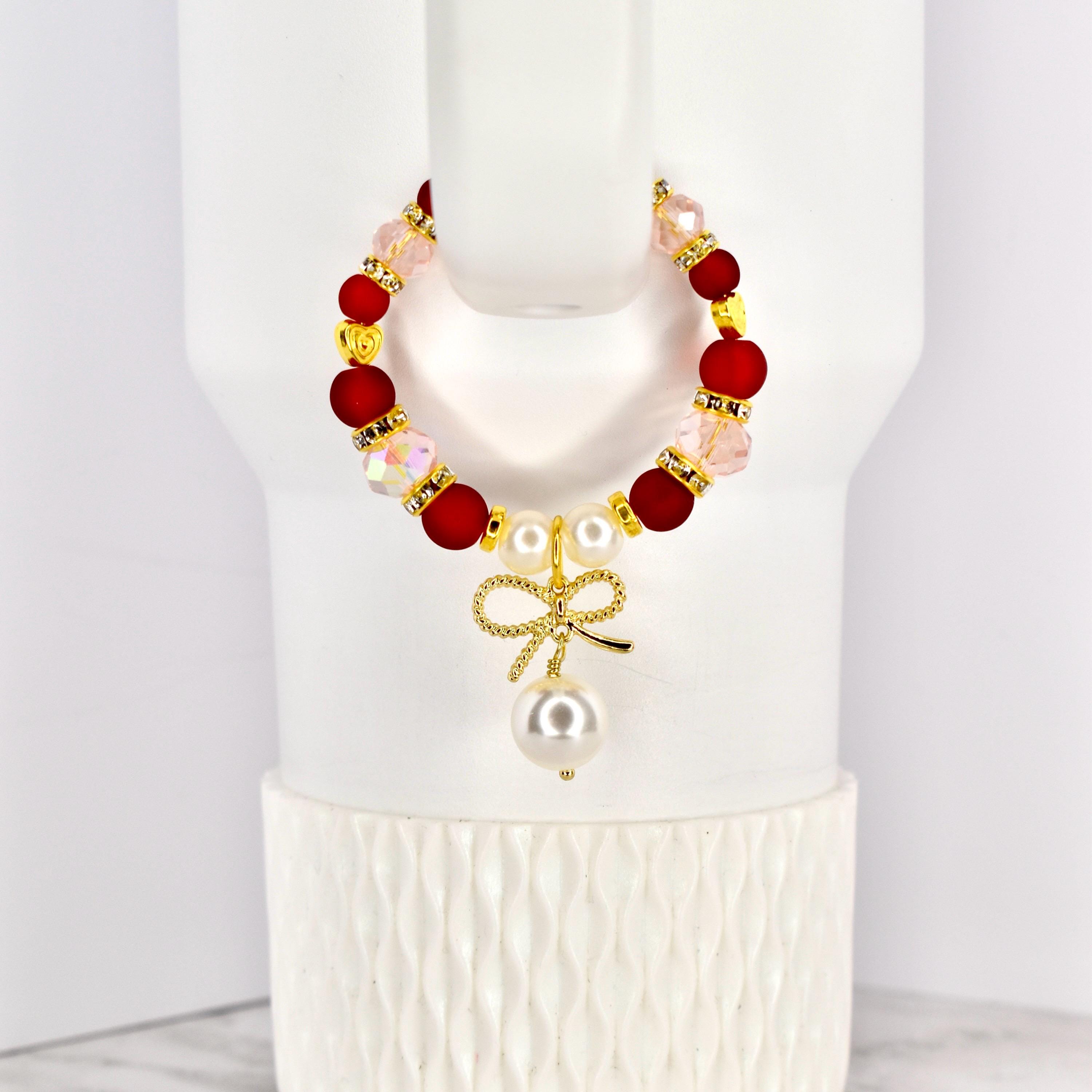A beaded tumbler handle bracelet with red, pink, and gold accents. The bracelet features a gold bow charm with a dangling pearl. The bracelet is displayed on a white Stanley.