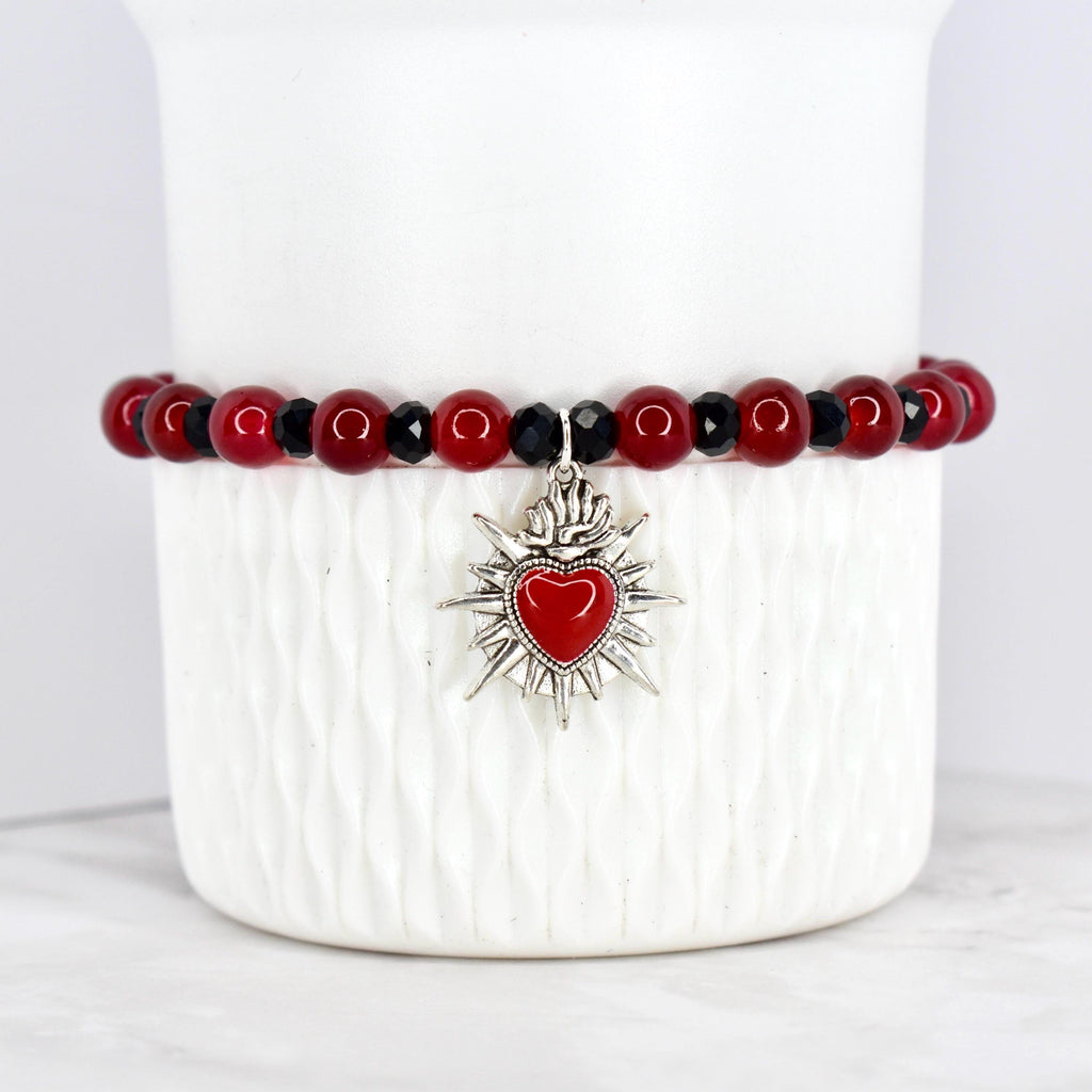A beaded bracelet featuring red and black beads. The bracelet has a silver-toned charm with a red heart at the center, surrounded by a sunburst design. The bracelet is displayed on a white, textured surface.