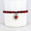 A beaded bracelet featuring red and black beads. The bracelet has a silver-toned charm with a red heart at the center, surrounded by a sunburst design. The bracelet is displayed on a white, textured surface.