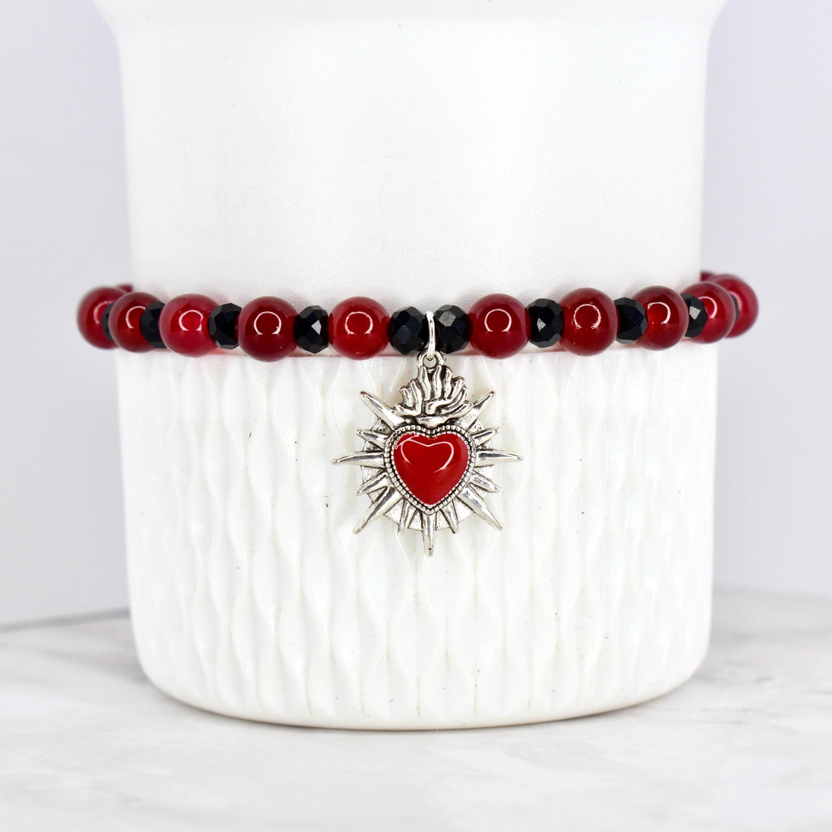 A beaded bracelet featuring red and black beads. The bracelet has a silver-toned charm with a red heart at the center, surrounded by a sunburst design. The bracelet is displayed on a white, textured surface.