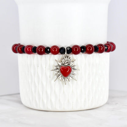 A beaded bracelet featuring red and black beads. The bracelet has a silver-toned charm with a red heart at the center, surrounded by a sunburst design. The bracelet is displayed on a white, textured surface.