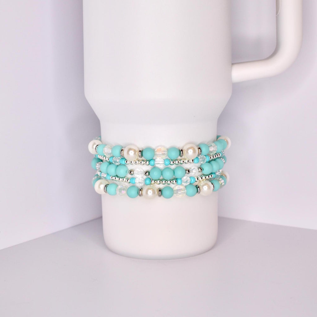 A white Stanley tumbler with a handle, shown with a set of five turquoise-themed beaded tumbler bracelets stacked around the base. The bracelets feature a mix of matte turquoise, iridescent crystal, pearl-like white, and shiny silver beads for a bright, summery look.