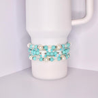 A white Stanley tumbler with a handle, shown with a set of five turquoise-themed beaded tumbler bracelets stacked around the base. The bracelets feature a mix of matte turquoise, iridescent crystal, pearl-like white, and shiny silver beads for a bright, summery look.