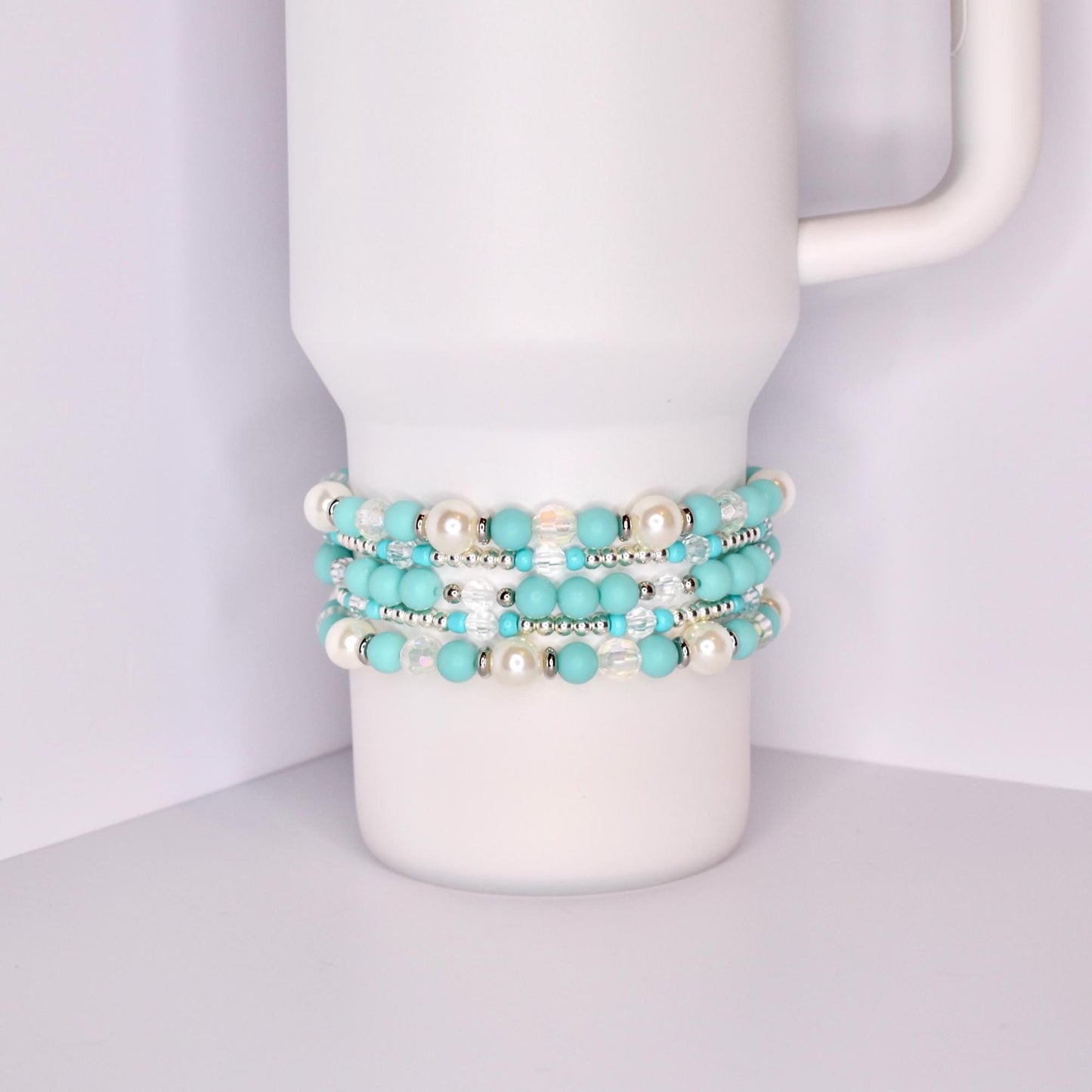 A white Stanley tumbler with a handle, shown with a set of five turquoise-themed beaded tumbler bracelets stacked around the base. The bracelets feature a mix of matte turquoise, iridescent crystal, pearl-like white, and shiny silver beads for a bright, summery look.