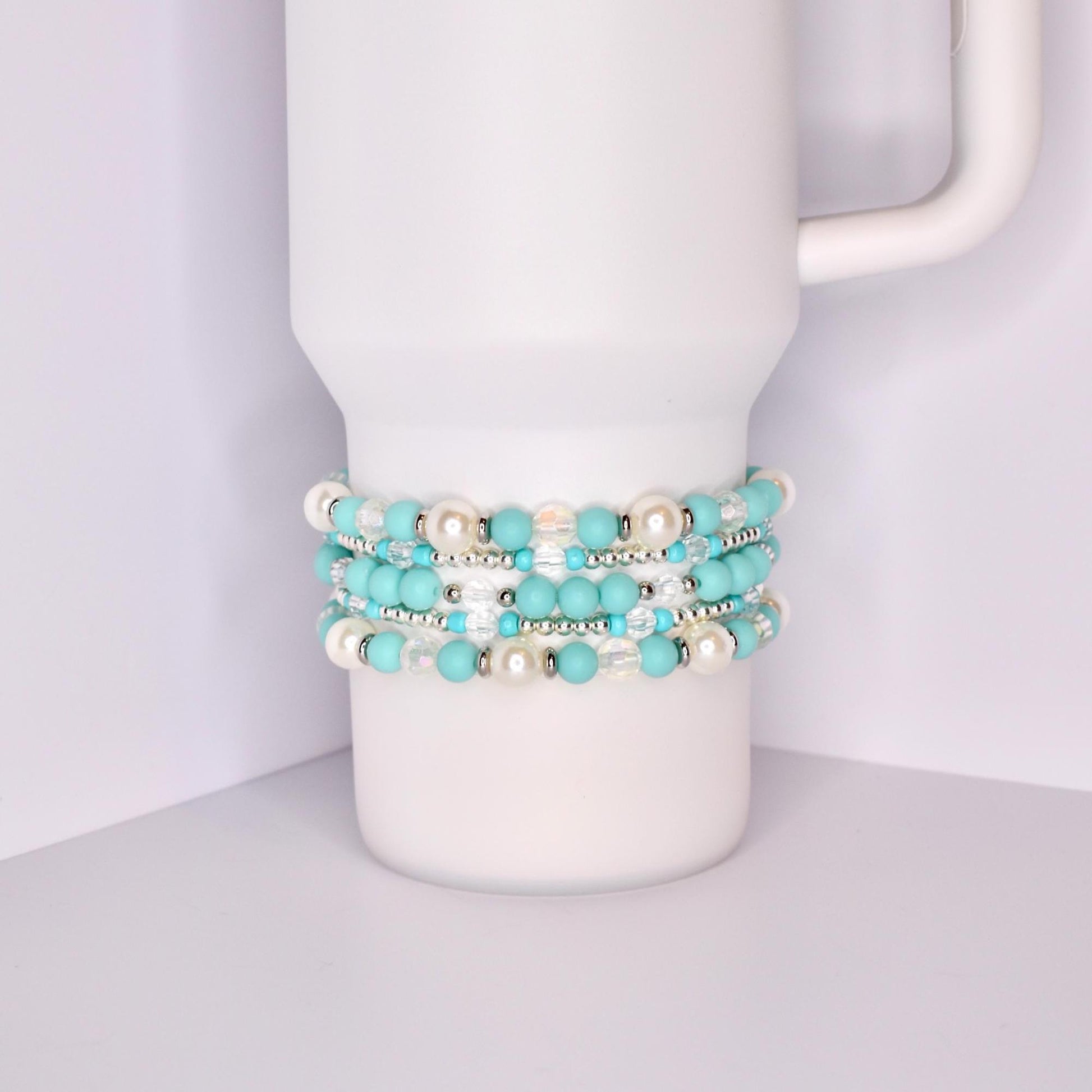 A white Stanley tumbler with a handle, shown with a set of five turquoise-themed beaded tumbler bracelets stacked around the base. The bracelets feature a mix of matte turquoise, iridescent crystal, pearl-like white, and shiny silver beads for a bright, summery look.