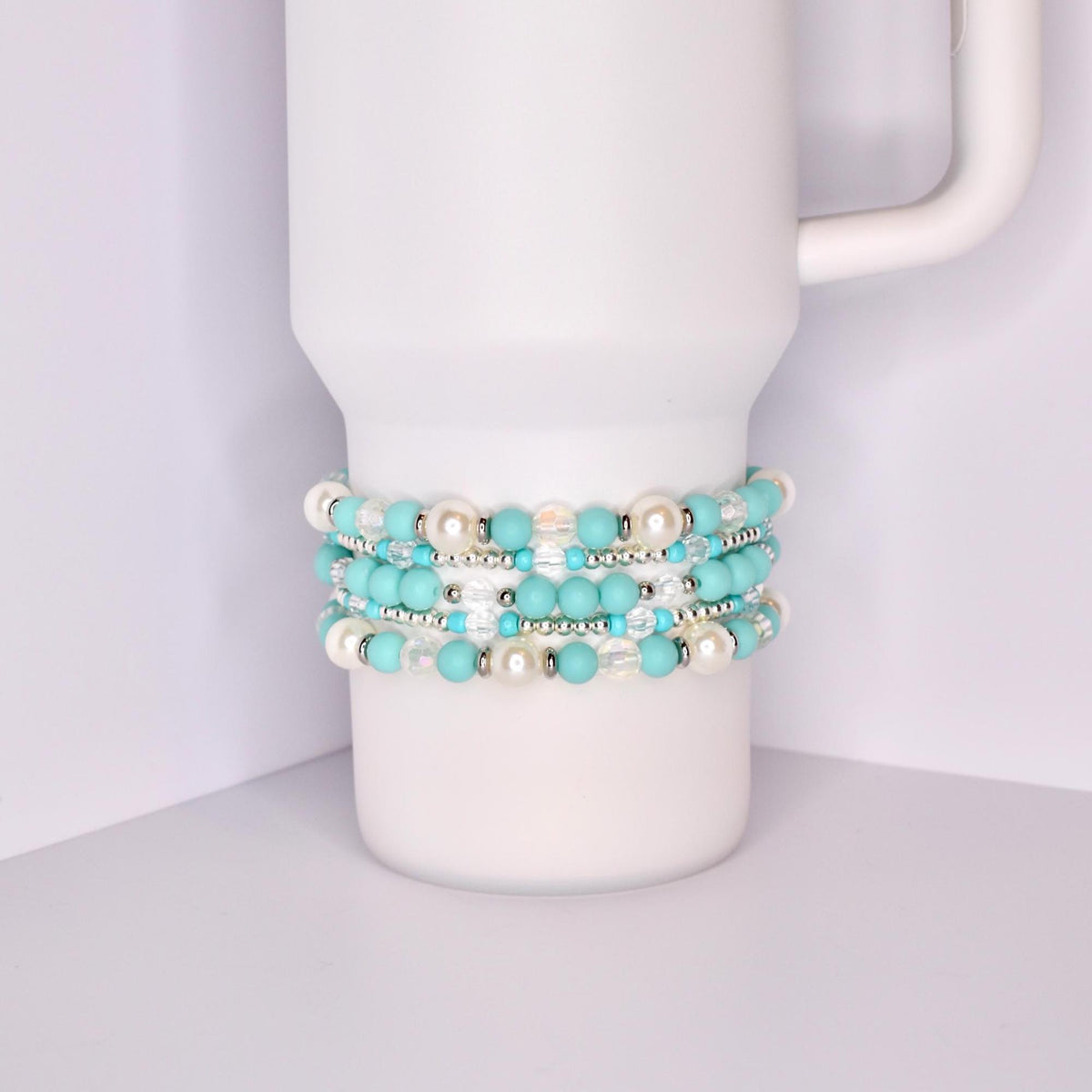 A white Stanley tumbler with a handle, shown with a set of five turquoise-themed beaded tumbler bracelets stacked around the base. The bracelets feature a mix of matte turquoise, iridescent crystal, pearl-like white, and shiny silver beads for a bright, summery look.