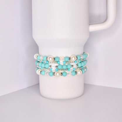 A white Stanley tumbler with a handle, shown with a set of five turquoise-themed beaded tumbler bracelets stacked around the base. The bracelets feature a mix of matte turquoise, iridescent crystal, pearl-like white, and shiny silver beads for a bright, summery look.
