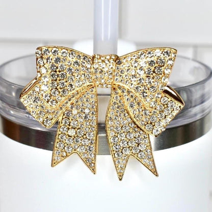 A gold-toned bow-shaped tumbler straw charm, encrusted with clear crystals, adorns a white tumbler. The bow is centered on the tumbler's rim, adding a touch of glamour. The background is a white tiled surface.
