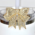 A gold-toned bow-shaped tumbler straw charm, encrusted with clear crystals, adorns a white tumbler. The bow is centered on the tumbler's rim, adding a touch of glamour. The background is a white tiled surface.