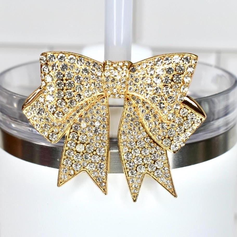 A gold-toned bow-shaped tumbler straw charm, encrusted with clear crystals, adorns a white tumbler. The bow is centered on the tumbler's rim, adding a touch of glamour. The background is a white tiled surface.