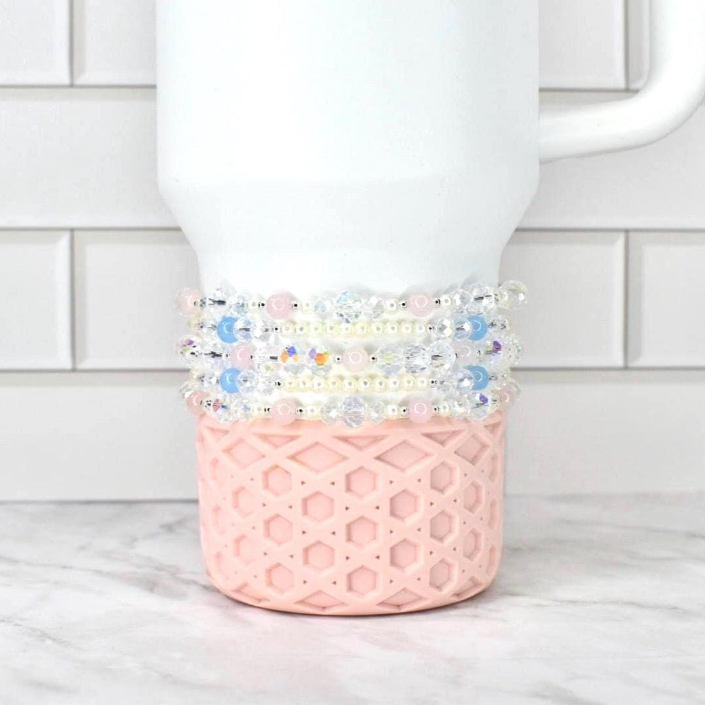 A white Stanley tumbler with a pink, geometric patterned base. Several beaded bracelets in shades of pink, blue, and clear are wrapped around the tumbler. The background is a white brick wall and a marble surface.