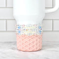 A white Stanley tumbler with a pink, geometric patterned base. Several beaded bracelets in shades of pink, blue, and clear are wrapped around the tumbler. The background is a white brick wall and a marble surface.