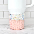 A white Stanley tumbler with a pink, geometric patterned base. Several beaded bracelets in shades of pink, blue, and clear are wrapped around the tumbler. The background is a white brick wall and a marble surface.