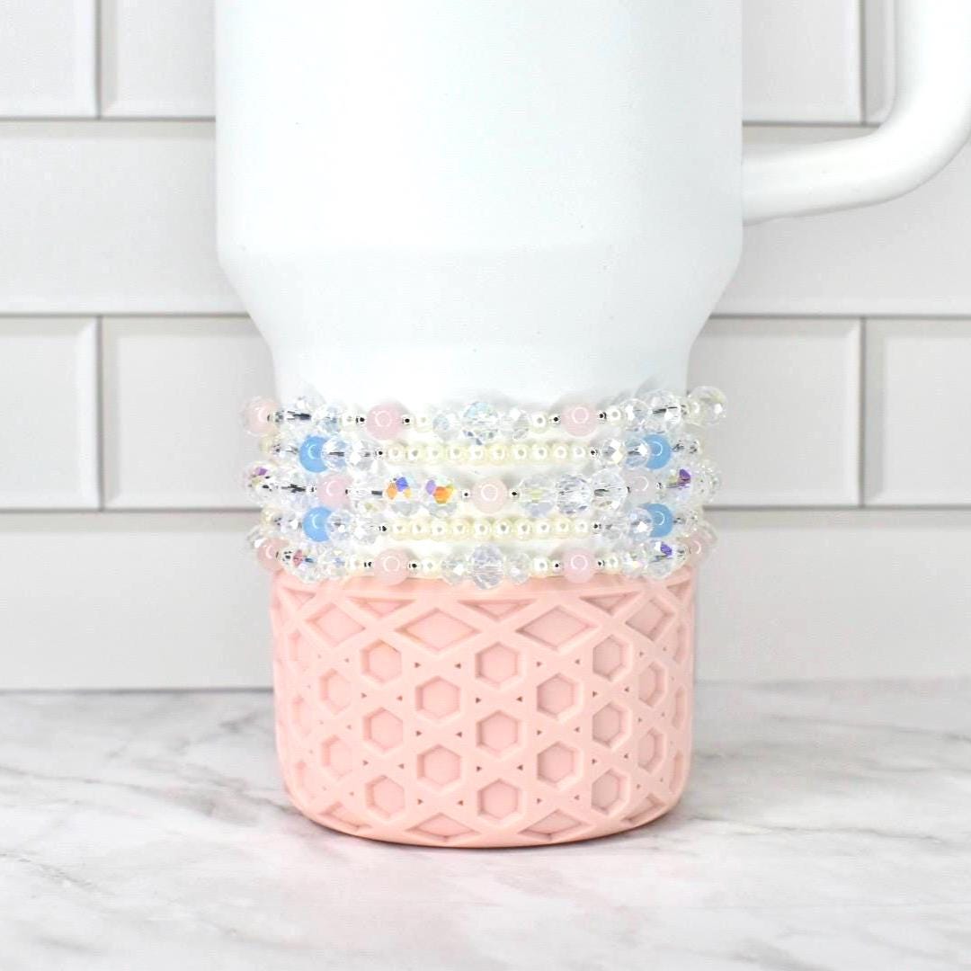 A white Stanley tumbler with a pink, geometric patterned base. Several beaded bracelets in shades of pink, blue, and clear are wrapped around the tumbler. The background is a white brick wall and a marble surface.