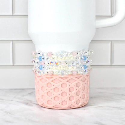 A white Stanley tumbler with a pink, geometric patterned base. Several beaded bracelets in shades of pink, blue, and clear are wrapped around the tumbler. The background is a white brick wall and a marble surface.