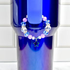 A beaded bracelet with blue, clear, and pink beads, and a star charm. The bracelet is displayed on a blue tumbler. The beads are faceted and round, with silver-colored spacers. The star charm is outlined in silver.