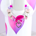 A beaded bracelet with pink, purple, and clear beads. The bracelet has a silver heart charm. The bracelet is displayed on a white surface with a pink and purple heart design.