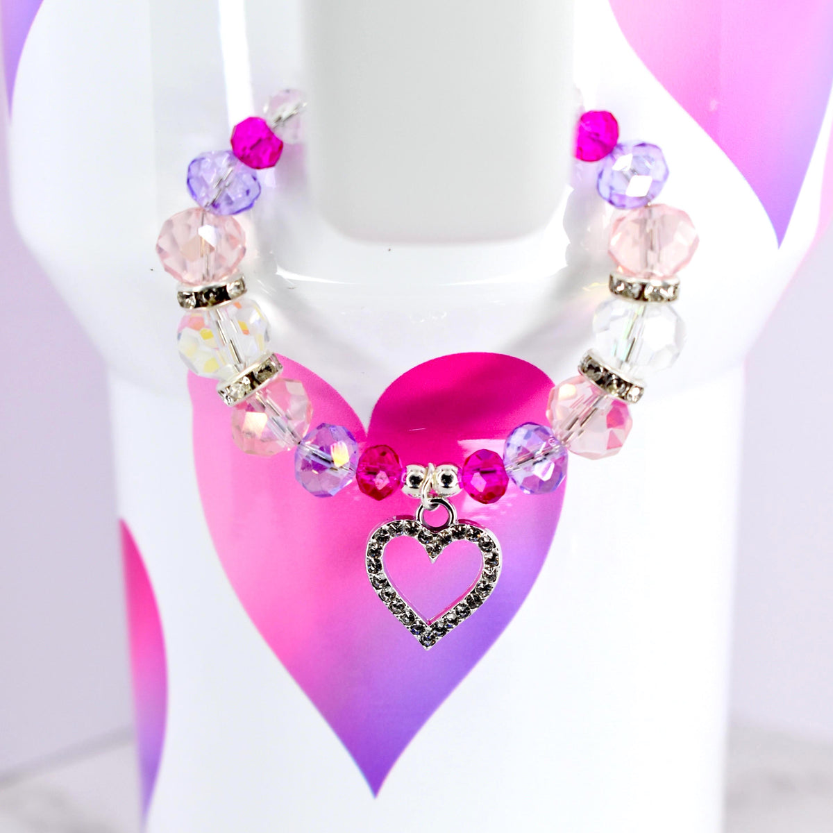 A beaded bracelet with pink, purple, and clear beads. The bracelet has a silver heart charm. The bracelet is displayed on a white surface with a pink and purple heart design.