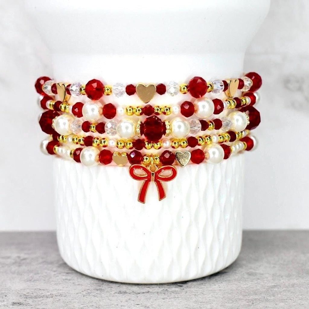 A stack of beaded bracelets in red, white, and gold. The bracelets feature various bead sizes and shapes, including round, faceted, and pearl-like beads. Gold heart charms and a red bow charm add detail.