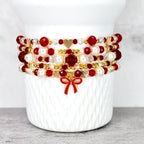 A stack of beaded bracelets in red, white, and gold. The bracelets feature various bead sizes and shapes, including round, faceted, and pearl-like beads. Gold heart charms and a red bow charm add detail.
