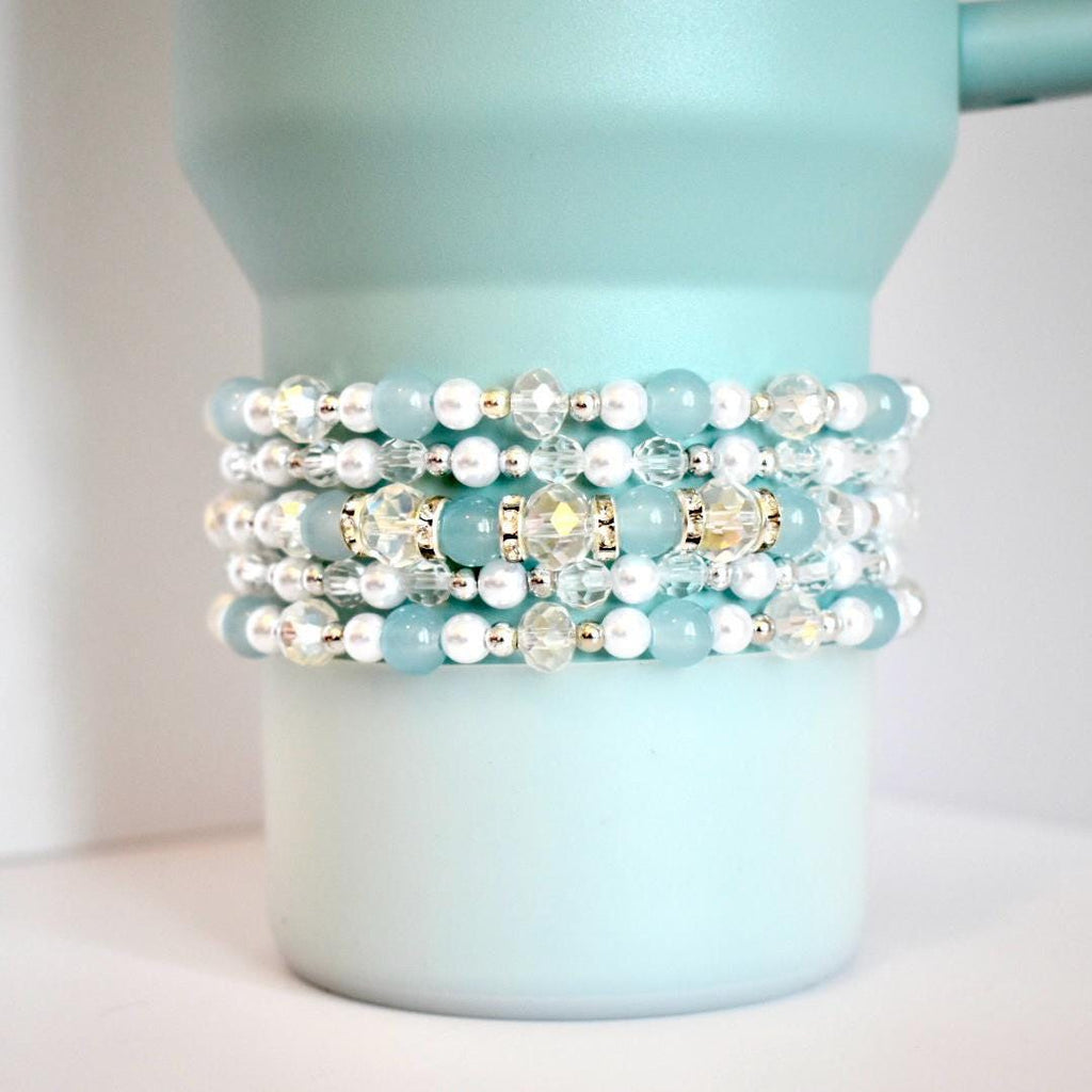 A stack of five beaded tumbler bracelets wrapped around a seafoam green Stanley-style tumbler. The bracelets feature seafoam green, pearl white, and clear crystal beads with metallic gold and silver accents, creating a sparkling and coastal-inspired look.