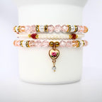 Three stacked bracelets with pink and white beads. The top and bottom bracelets have clear, faceted beads with gold accents and white pearl-like beads. The middle bracelet is made of small white beads and red beads. The bottom bracelet has a heart-shaped charm with a rose design and a dangling clear gem.