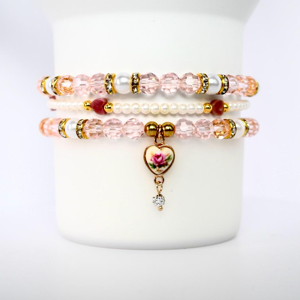 Three stacked bracelets with pink and white beads. The top and bottom bracelets have clear, faceted beads with gold accents and white pearl-like beads. The middle bracelet is made of small white beads and red beads. The bottom bracelet has a heart-shaped charm with a rose design and a dangling clear gem.
