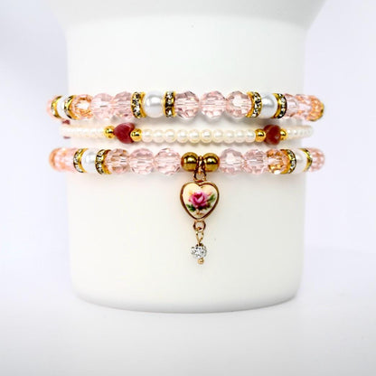 Three stacked bracelets with pink and white beads. The top and bottom bracelets have clear, faceted beads with gold accents and white pearl-like beads. The middle bracelet is made of small white beads and red beads. The bottom bracelet has a heart-shaped charm with a rose design and a dangling clear gem.