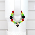 A colorful beaded tumbler handle bracelet featuring red, green, yellow, and black beads. The bracelet has a gold-toned heart charm with a black center. The bracelet is displayed on a white background.