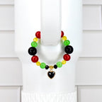 A colorful beaded tumbler handle bracelet featuring red, green, yellow, and black beads. The bracelet has a gold-toned heart charm with a black center. The bracelet is displayed on a white background.