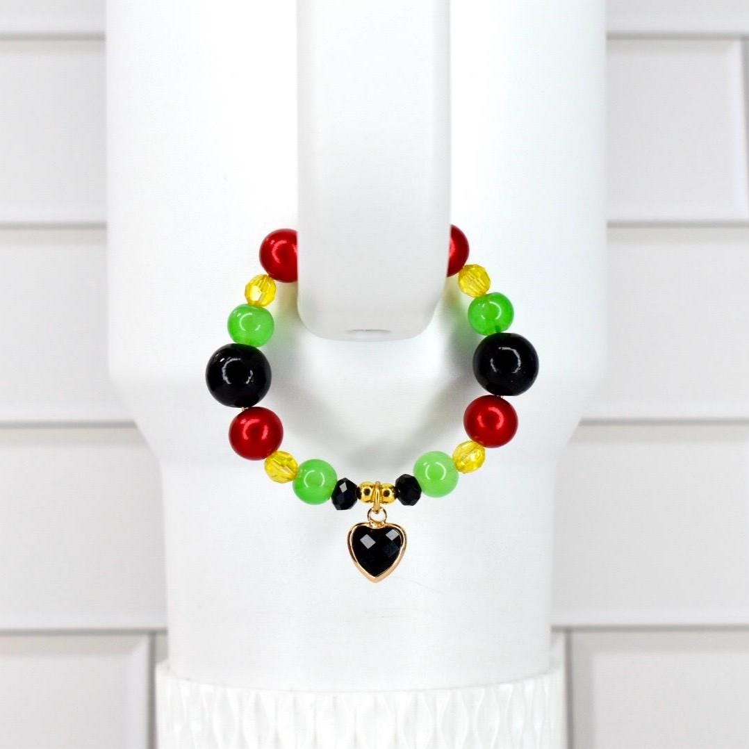 A colorful beaded tumbler handle bracelet featuring red, green, yellow, and black beads. The bracelet has a gold-toned heart charm with a black center. The bracelet is displayed on a white background.