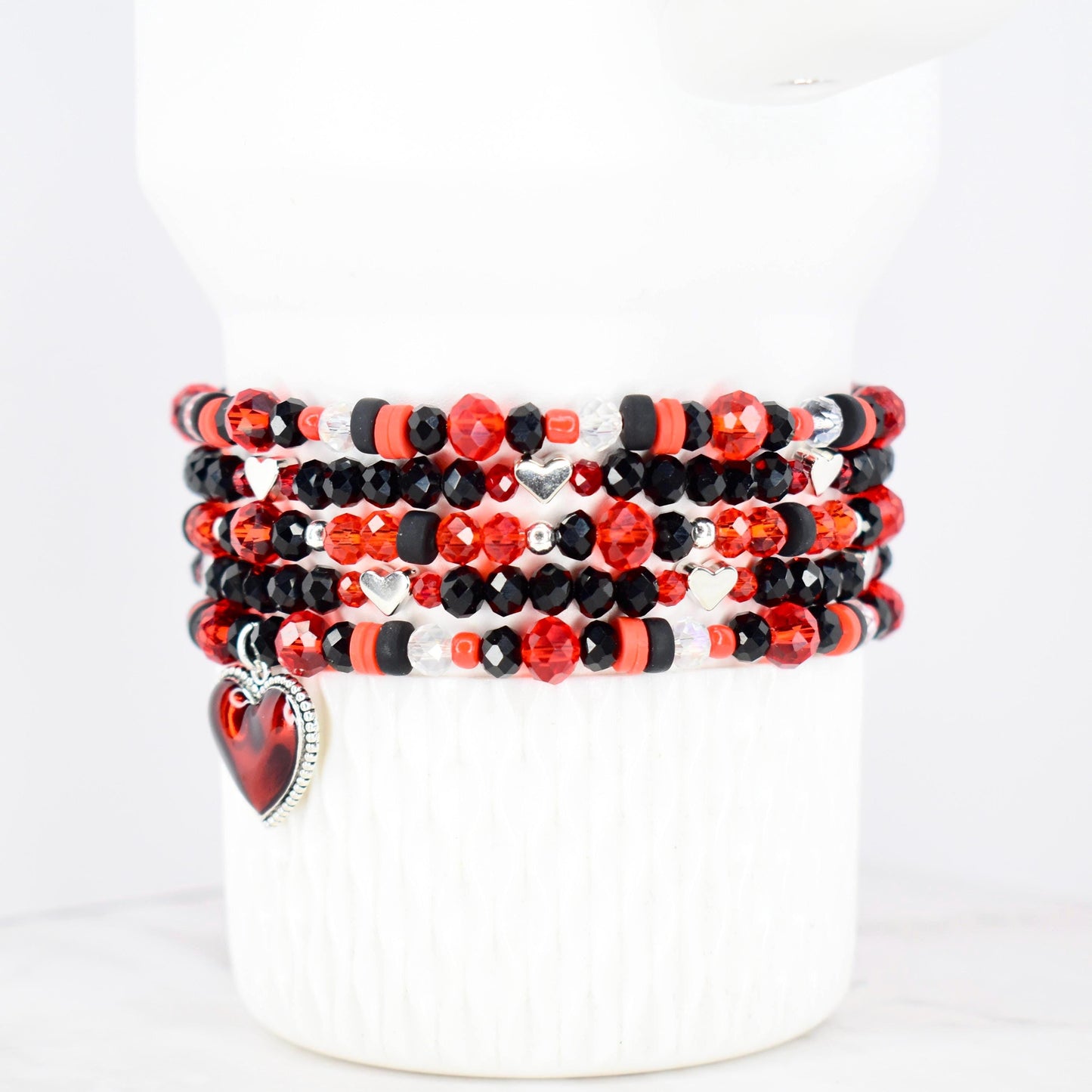 Stack of beaded bracelets in red, black, and clear beads with silver heart charms. The bracelets are displayed on a white cylindrical form. The beads are various shapes and sizes, with a heart-shaped charm dangling from one bracelet.