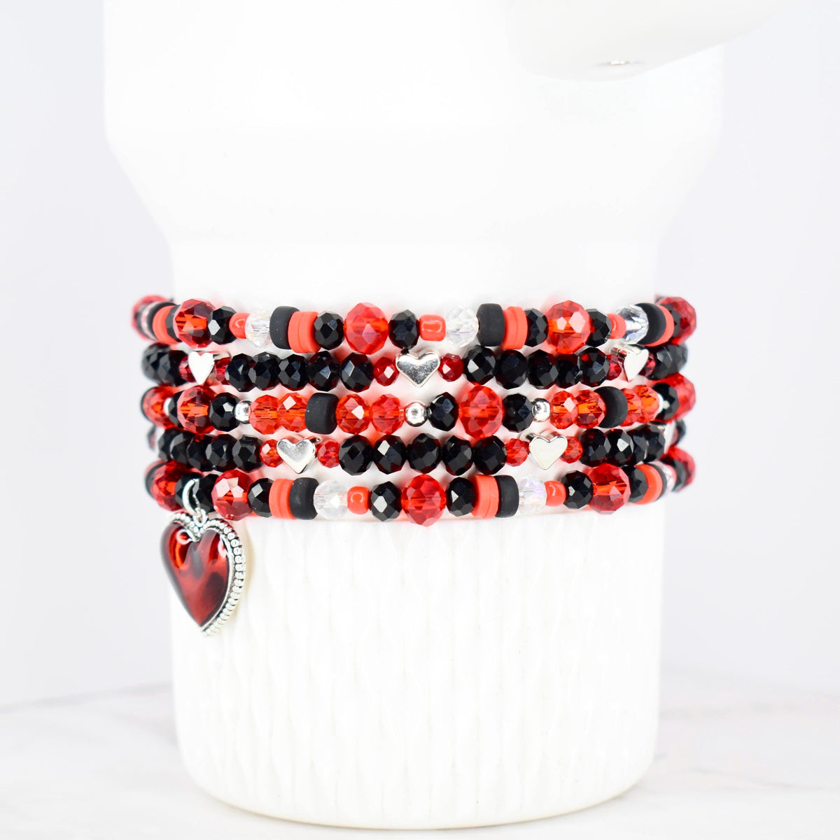 Stack of beaded bracelets in red, black, and clear beads with silver heart charms. The bracelets are displayed on a white cylindrical form. The beads are various shapes and sizes, with a heart-shaped charm dangling from one bracelet.
