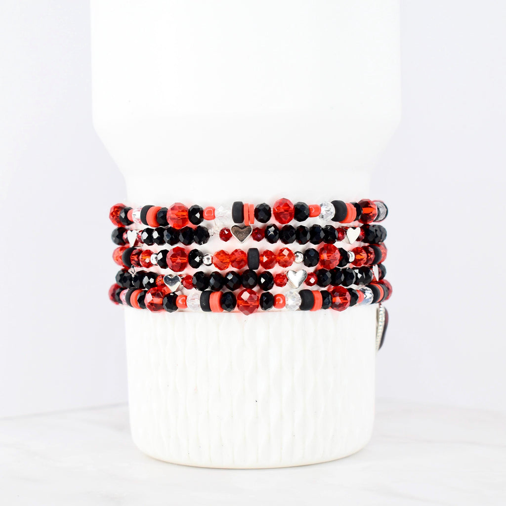 Stack of beaded bracelets in red, black, and silver. The bracelets feature various bead shapes and sizes, including faceted beads and small heart charms. The bracelets are displayed on a white cylindrical object.