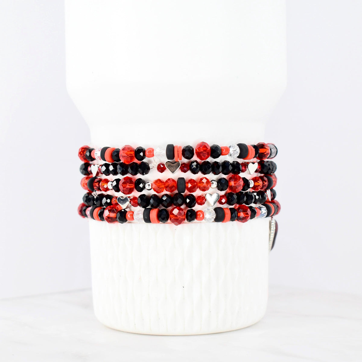 Stack of beaded bracelets in red, black, and silver. The bracelets feature various bead shapes and sizes, including faceted beads and small heart charms. The bracelets are displayed on a white cylindrical object.