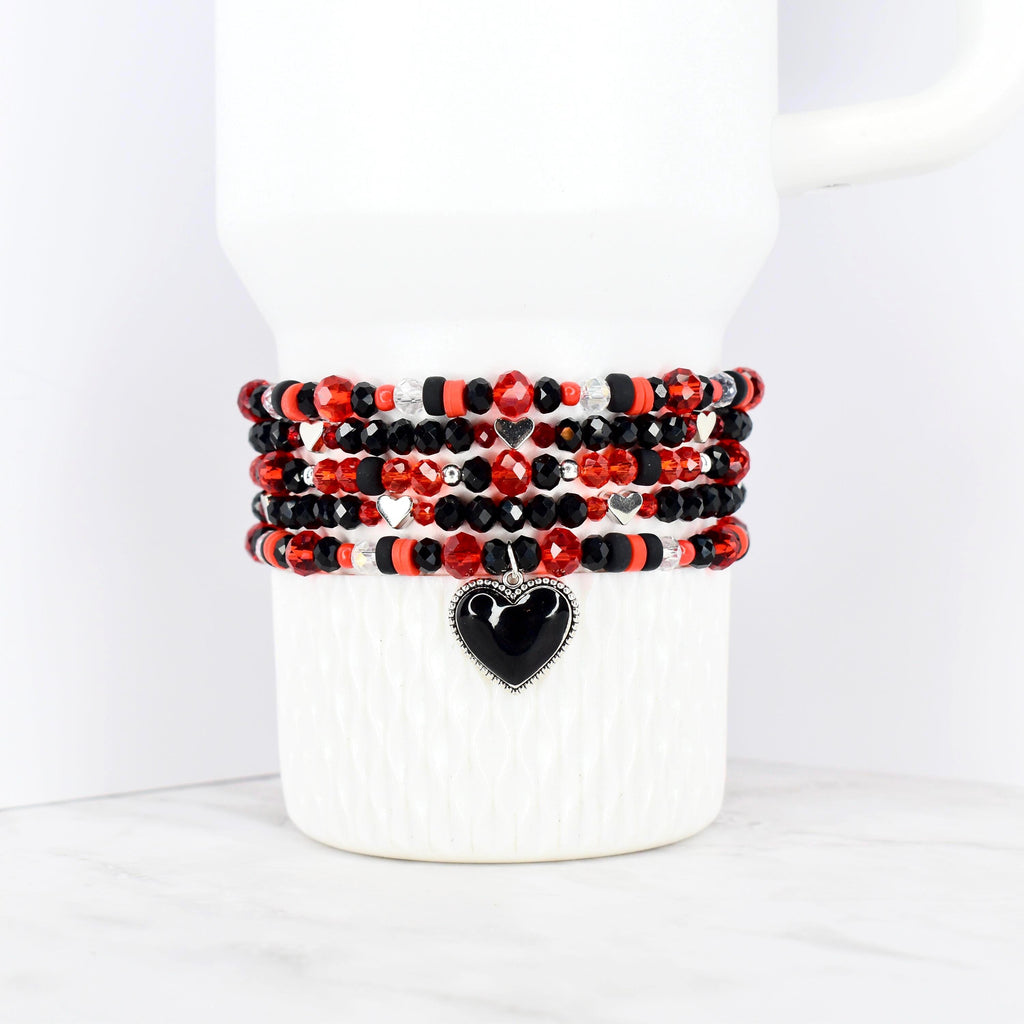 Stack of beaded bracelets in red, black, and silver, wrapped around a white mug. Each bracelet features a mix of beads, with a black heart charm on the bottom bracelet. The bracelets are a mix of faceted and round beads.