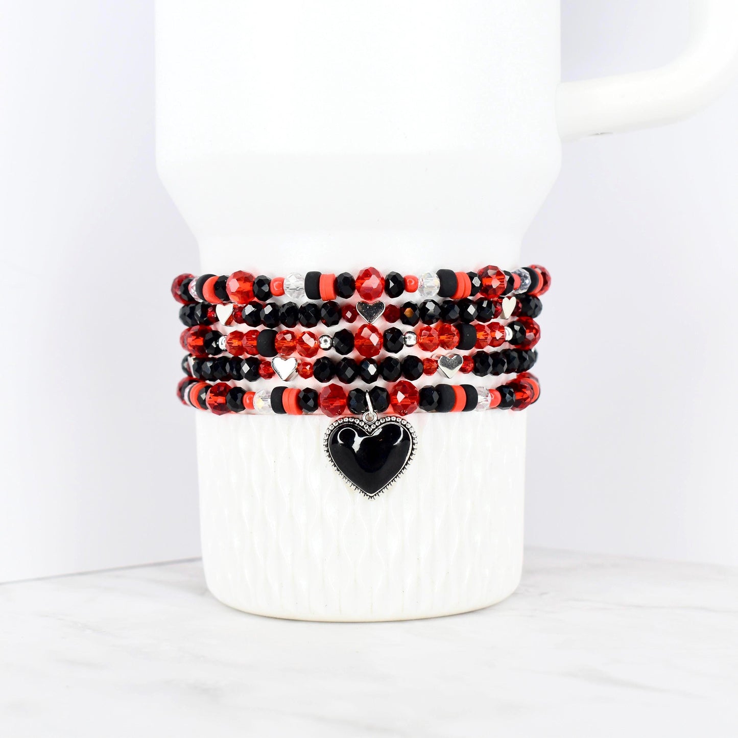 Stack of beaded bracelets in red, black, and silver, wrapped around a white mug. Each bracelet features a mix of beads, with a black heart charm on the bottom bracelet. The bracelets are a mix of faceted and round beads.