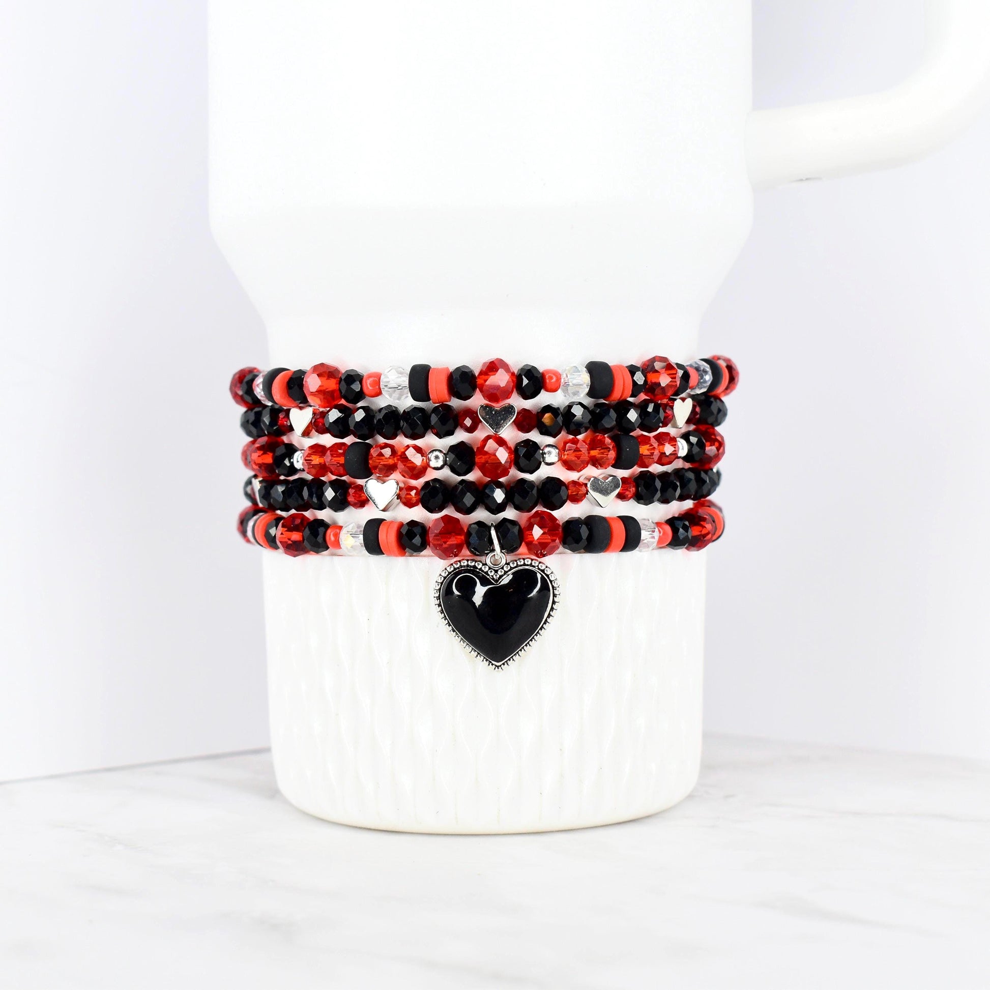 Stack of beaded bracelets in red, black, and silver, wrapped around a white mug. Each bracelet features a mix of beads, with a black heart charm on the bottom bracelet. The bracelets are a mix of faceted and round beads.