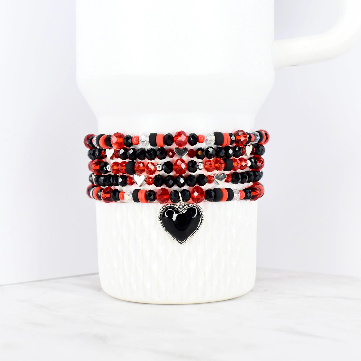 Stack of beaded bracelets in red, black, and silver, wrapped around a white mug. Each bracelet features a mix of beads, with a black heart charm on the bottom bracelet. The bracelets are a mix of faceted and round beads.