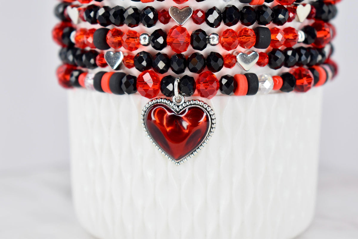 Stack of beaded bracelets in red, black, and silver. The bracelets feature various bead shapes and sizes, including faceted and round beads. A red heart pendant with a silver border hangs from the bottom bracelet.