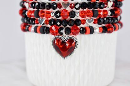 Stack of beaded bracelets in red, black, and silver. The bracelets feature various bead shapes and sizes, including faceted and round beads. A red heart pendant with a silver border hangs from the bottom bracelet.