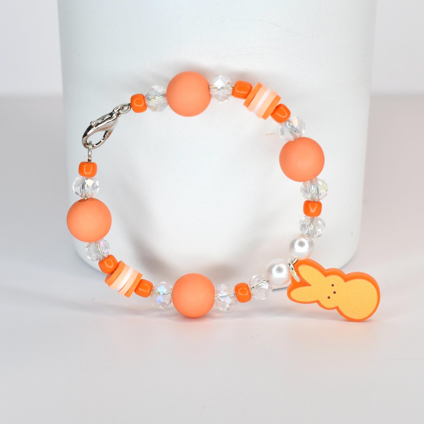A beaded bracelet featuring orange matte beads, clear crystal beads, and small orange and white striped beads. A charm in the shape of a yellow bunny is attached. The bracelet has a silver clasp.
