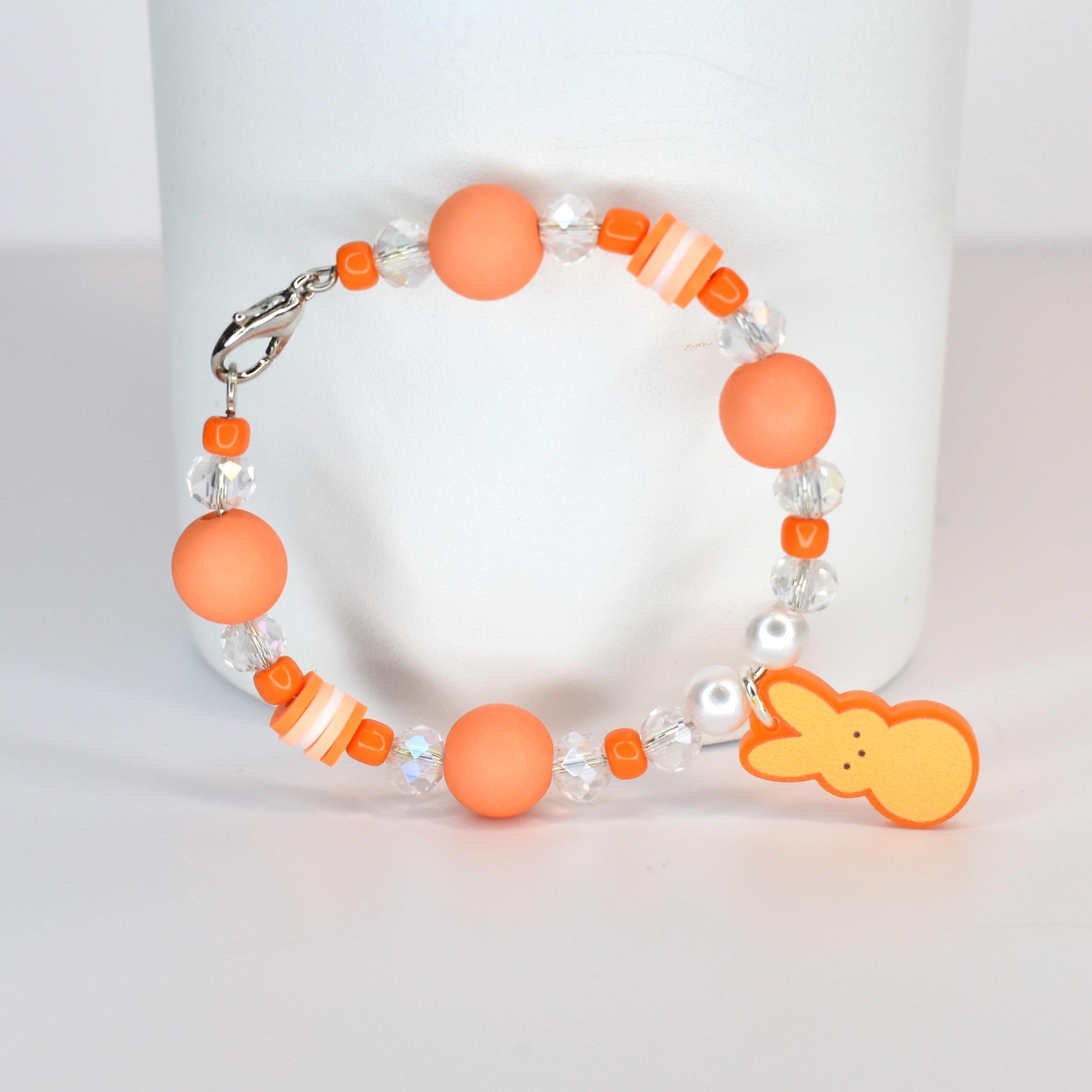 A beaded bracelet featuring orange matte beads, clear crystal beads, and small orange and white striped beads. A charm in the shape of a yellow bunny is attached. The bracelet has a silver clasp.