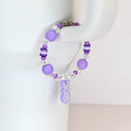 A purple beaded bracelet with a bunny charm. The bracelet features large round purple beads, clear crystal beads, and small white beads. The bunny charm is a light purple color. The bracelet is held up by a white surface.