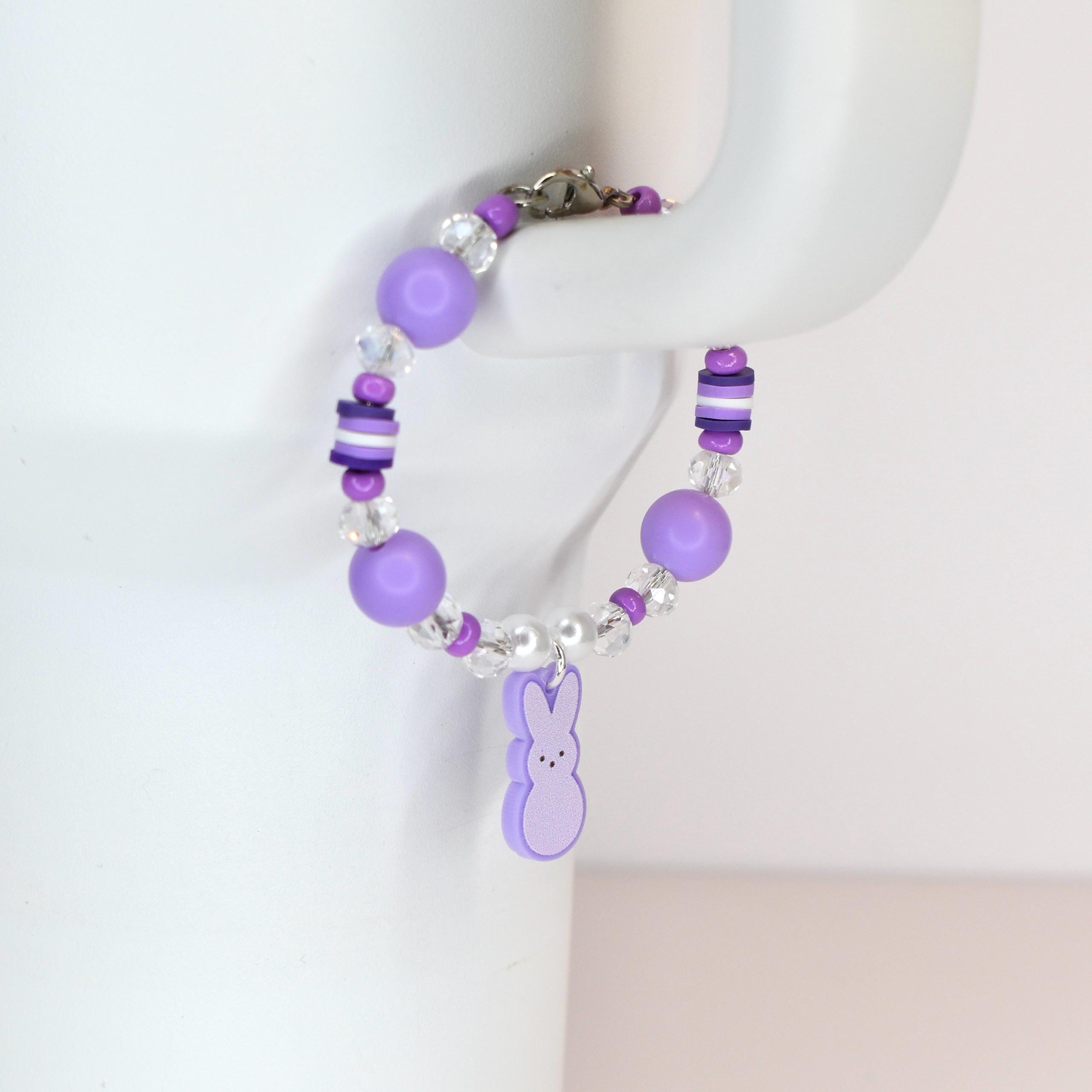 A purple beaded bracelet with a bunny charm. The bracelet features large round purple beads, clear crystal beads, and small white beads. The bunny charm is a light purple color. The bracelet is held up by a white surface.