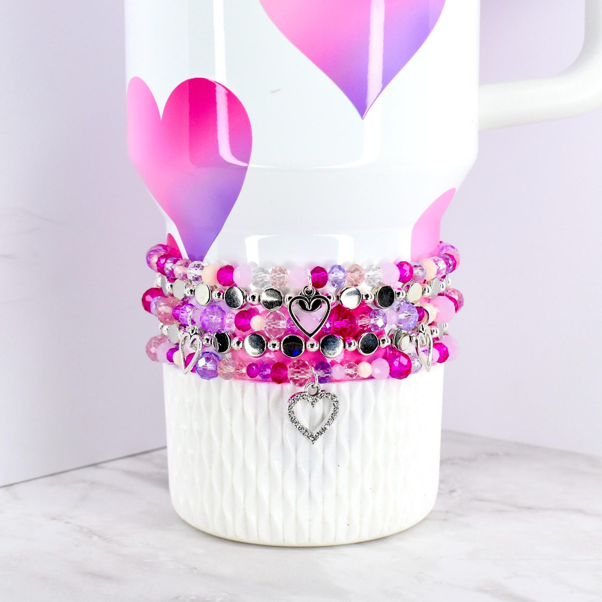 Stack of beaded bracelets in shades of pink, purple, and silver, with heart-shaped charms. The bracelets are displayed on a white cup with pink and purple heart designs. The beads vary in size and texture.