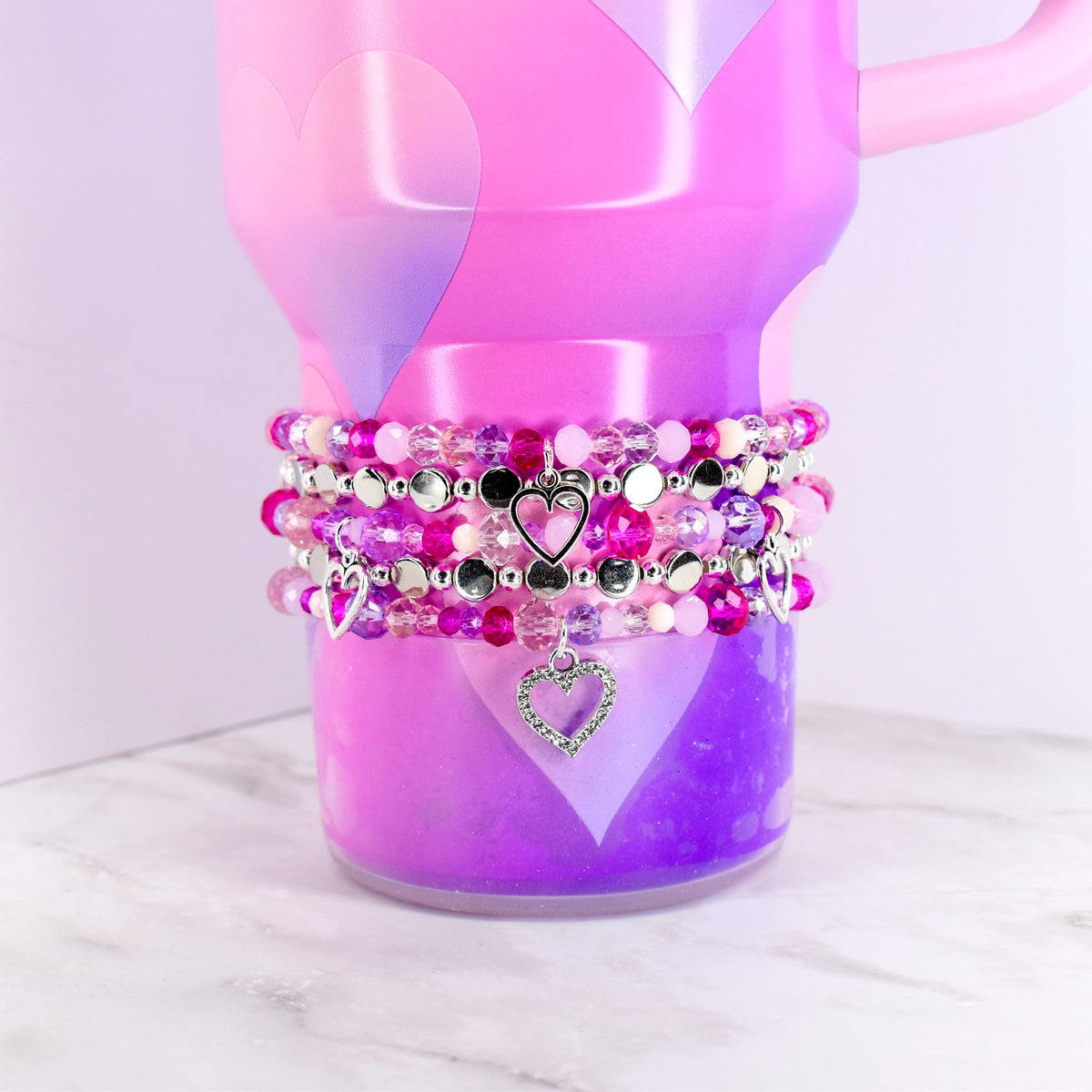 A pink and purple ombre tumbler with a handle, adorned with multiple beaded bracelets. The bracelets feature various shades of pink and purple beads, silver accents, and heart-shaped charms. The tumbler has heart-shaped designs.