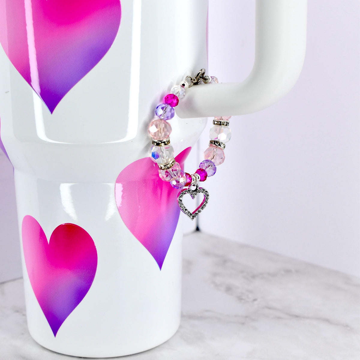 A beaded bracelet with pink, purple, and clear beads, and a silver heart charm. The bracelet is displayed on the handle of a white tumbler decorated with pink and purple heart designs.
